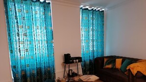 A living room with a brown sofa, a small table, and a coffee machine. There are curtains with a blue pattern and a wooden floor. - B&B 43 4 (Le Puy-en-Velay)