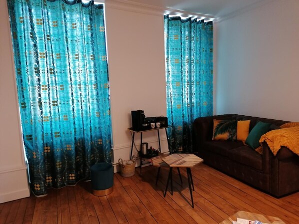 A living room with a brown sofa, a small table, and a coffee machine. There are curtains with a blue pattern and a wooden floor. - B&B 43 4 (Le Puy-en-Velay)