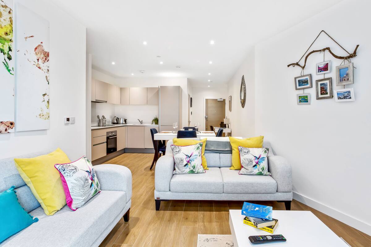 Una sala de estar moderna con un sofá gris, cojines coloridos, una mesa de centro con libros y un control remoto, y una cocina al fondo.