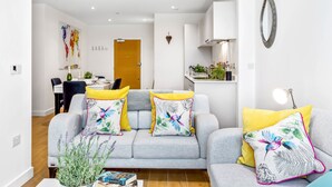 Una sala de estar moderna con un sofá gris, cojines amarillos y estampados florales, una pequeña mesa con un libro y una planta en maceta.