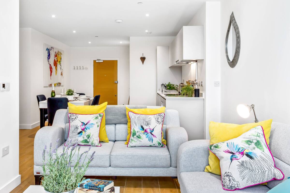 Una sala de estar moderna con un sofá gris, cojines amarillos y estampados florales, una pequeña mesa con un libro y una planta en maceta.