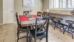 Un comedor con una mesa de madera, individuales rojos y sillas negras. Hay una ventana con persianas, un cuadro en la pared y una puerta.
