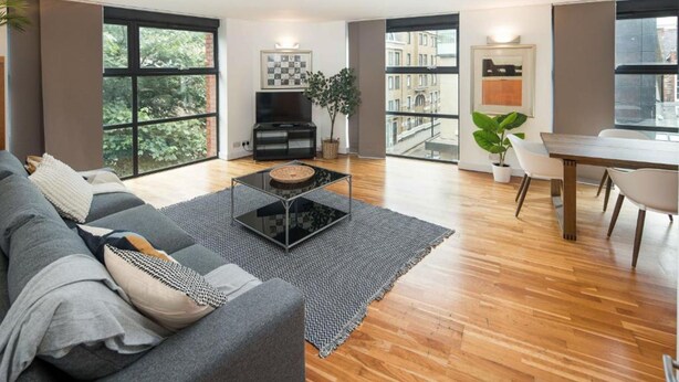 A modern living room with a grey sofa, a coffee table, and a dining area with a white table and chairs. There are large windows with a view of buildings and trees. - Broadband 2BD City Flat Farringdon Expedia (London)