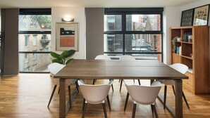 A modern dining area with a wooden table and white chairs, large windows with a view of buildings, and a bookshelf with books and plants. - Broadband 2BD City Flat Farringdon Expedia (London)