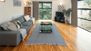 A modern living room with a grey sofa, a glass coffee table, and a large window offering a view of trees and buildings. - Broadband 2BD City Flat Farringdon Expedia (London)