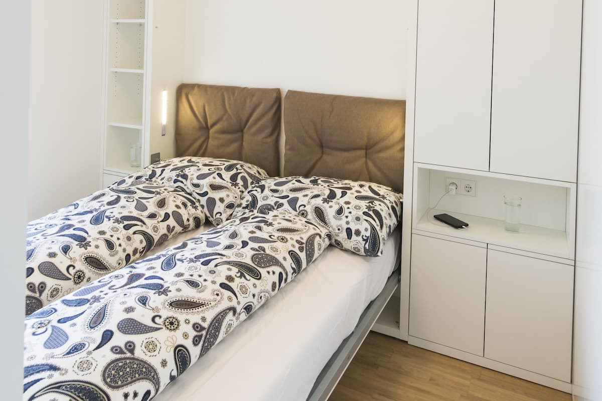 A bedroom with a bed, a headboard, a wardrobe, and a shelf with a remote control.