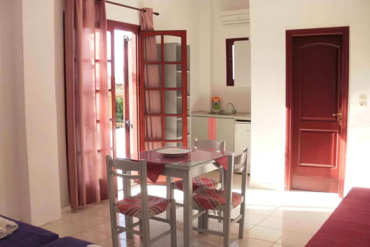 A room with a dining table, chairs, a bed, and a kitchen area.