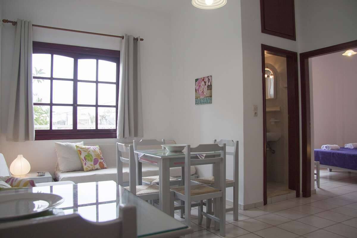 A room with a dining table and chairs, a window with curtains, a small table with a lamp, and a bed with a blue cover.