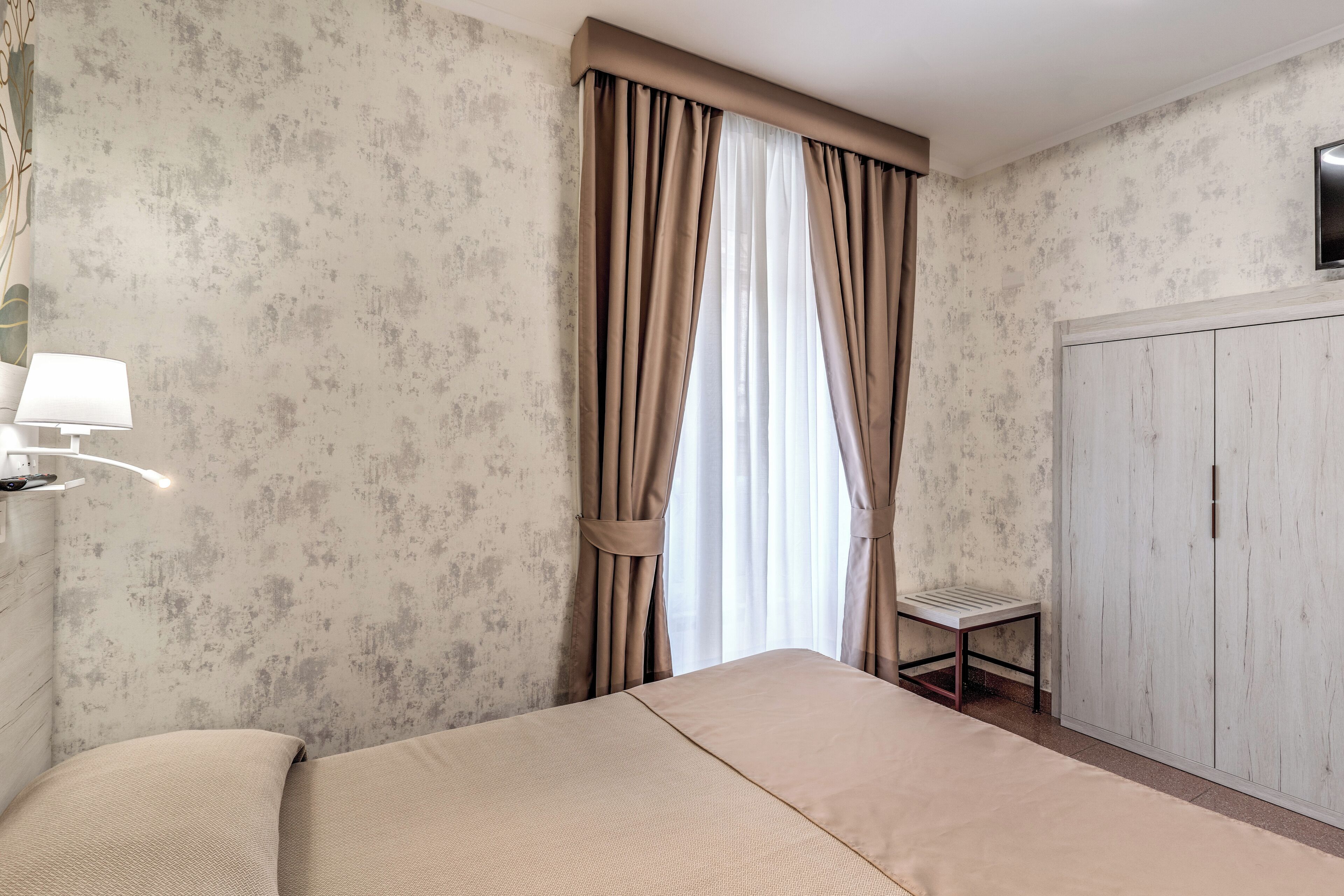 A hotel room with a bed, a small table, a lamp, and a wardrobe.