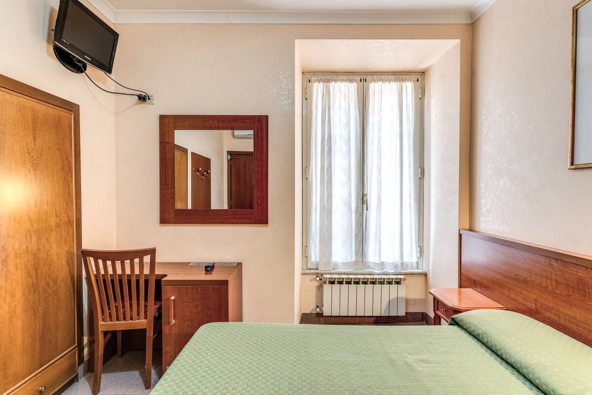 A hotel room with a bed, a desk, a chair, a television mounted on the wall, and a window with curtains.