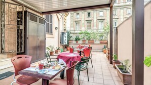 Un café en plein air avec des tables et des chaises, des plantes en pot et une vue sur un bâtiment en arrière-plan.