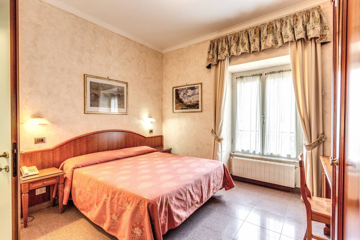 A hotel room with a large bed, two bedside tables, a nightstand, a window with curtains, and two wall-mounted lamps.