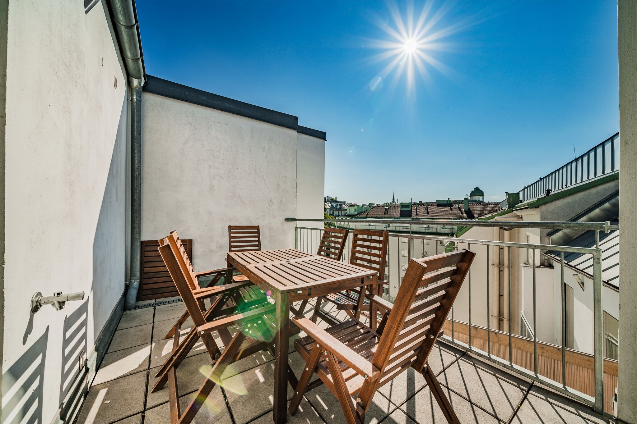 Un balcon extérieur avec une table et des chaises, une vue sur des bâtiments en arrière-plan et un ciel bleu dégagé.