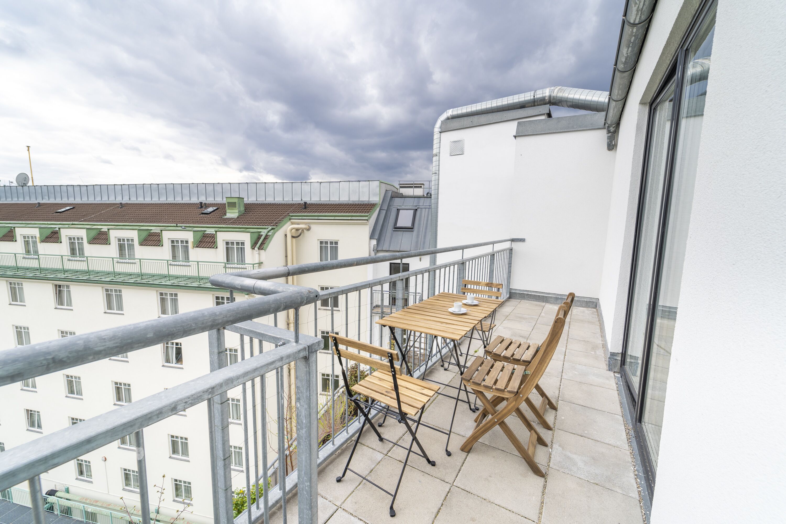 A balcony with a table and chairs, a metal railing, and a view of buildings in the background.
