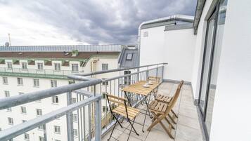 A balcony with a table and chairs, a metal railing, and a view of buildings in the background.