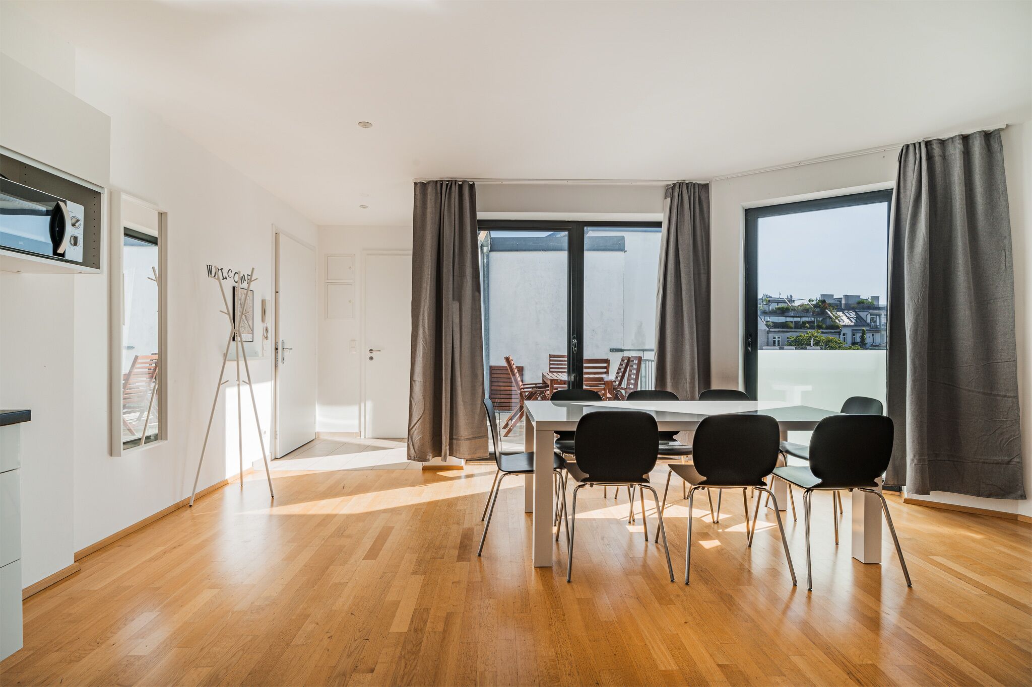 A modern dining area with a large table and chairs, large windows with curtains, and a view of buildings outside.