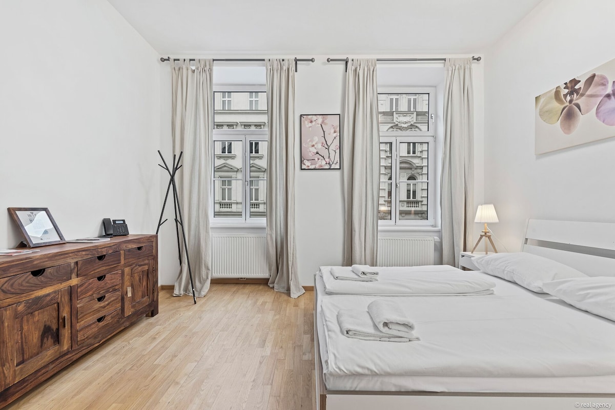 A bedroom with a wooden dresser, a bed with white linens, a standing lamp, and a view of buildings through the window.