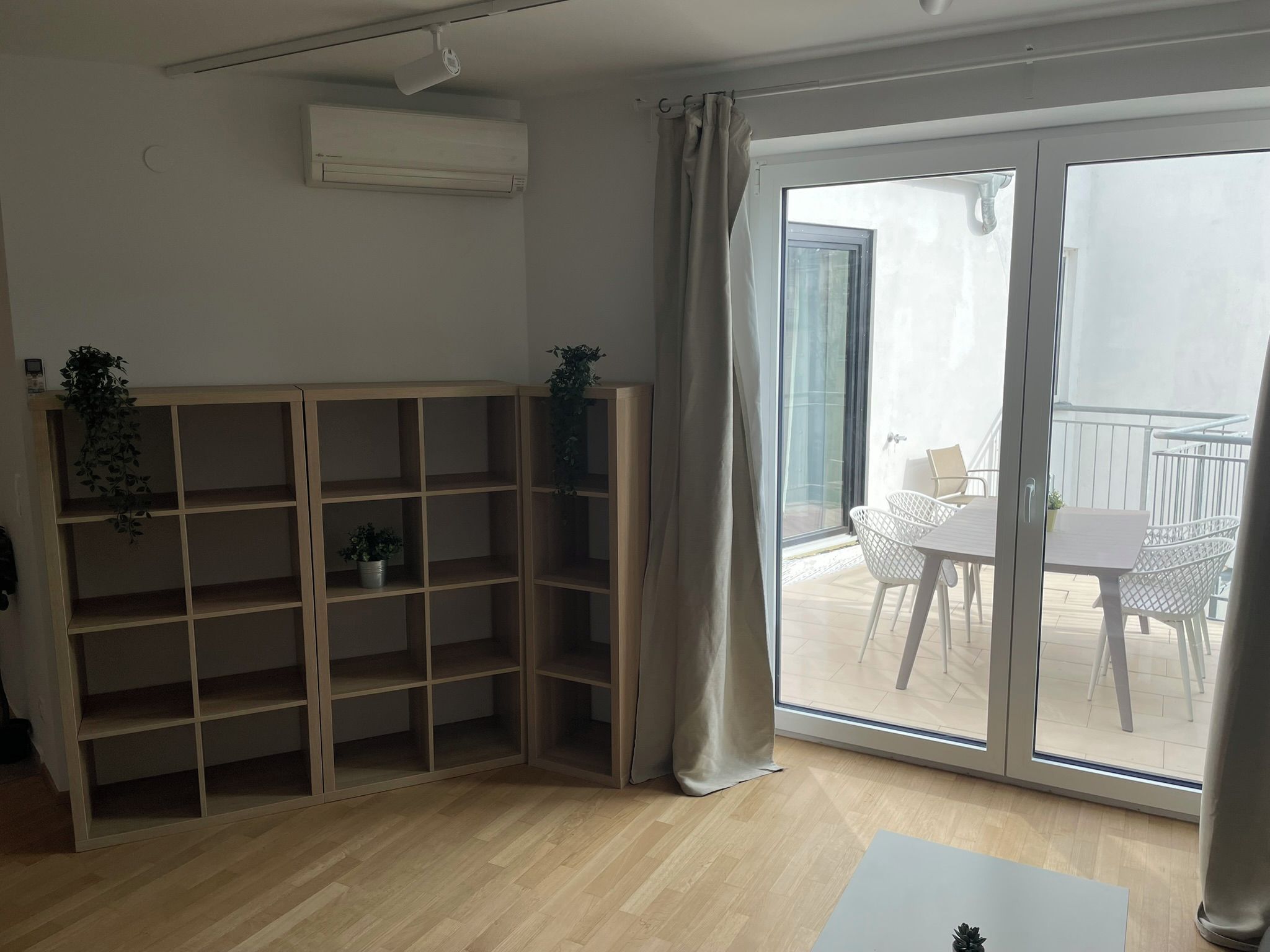 Ein Zimmer mit einer Holzbücherreol, einer Schiebetür zu einem Außenbereich mit Tisch und Stühlen sowie einer Klimaanlage an der Wand.