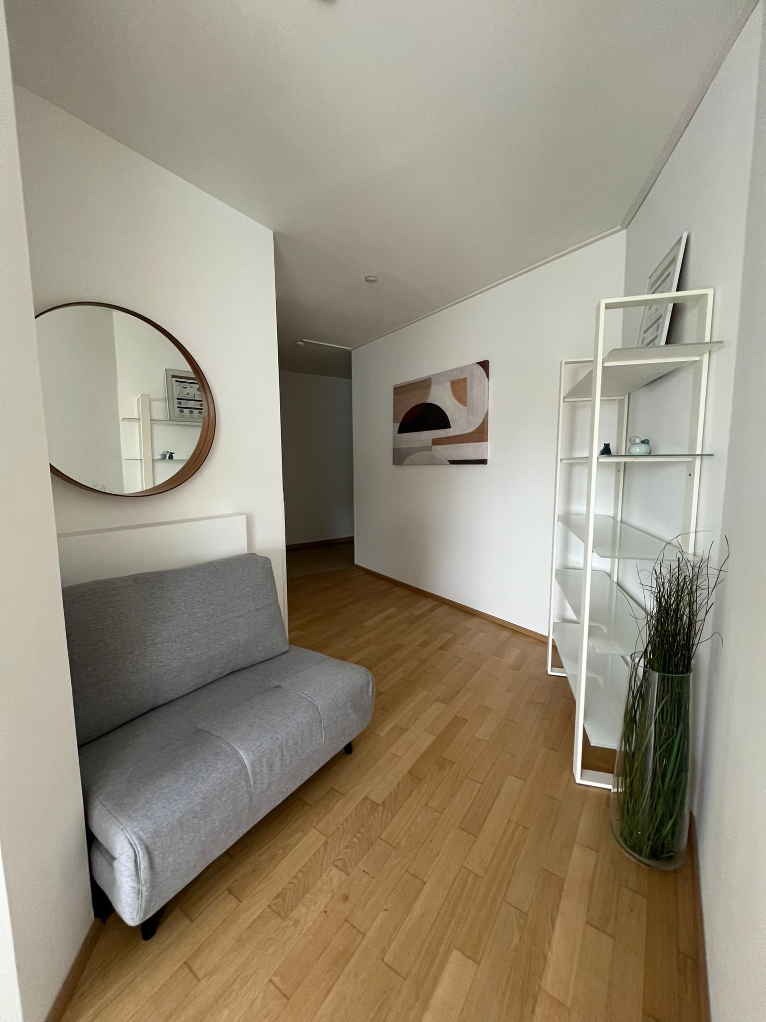 A modern living room with a grey sofa, a white shelving unit, and a mirror on the wall.