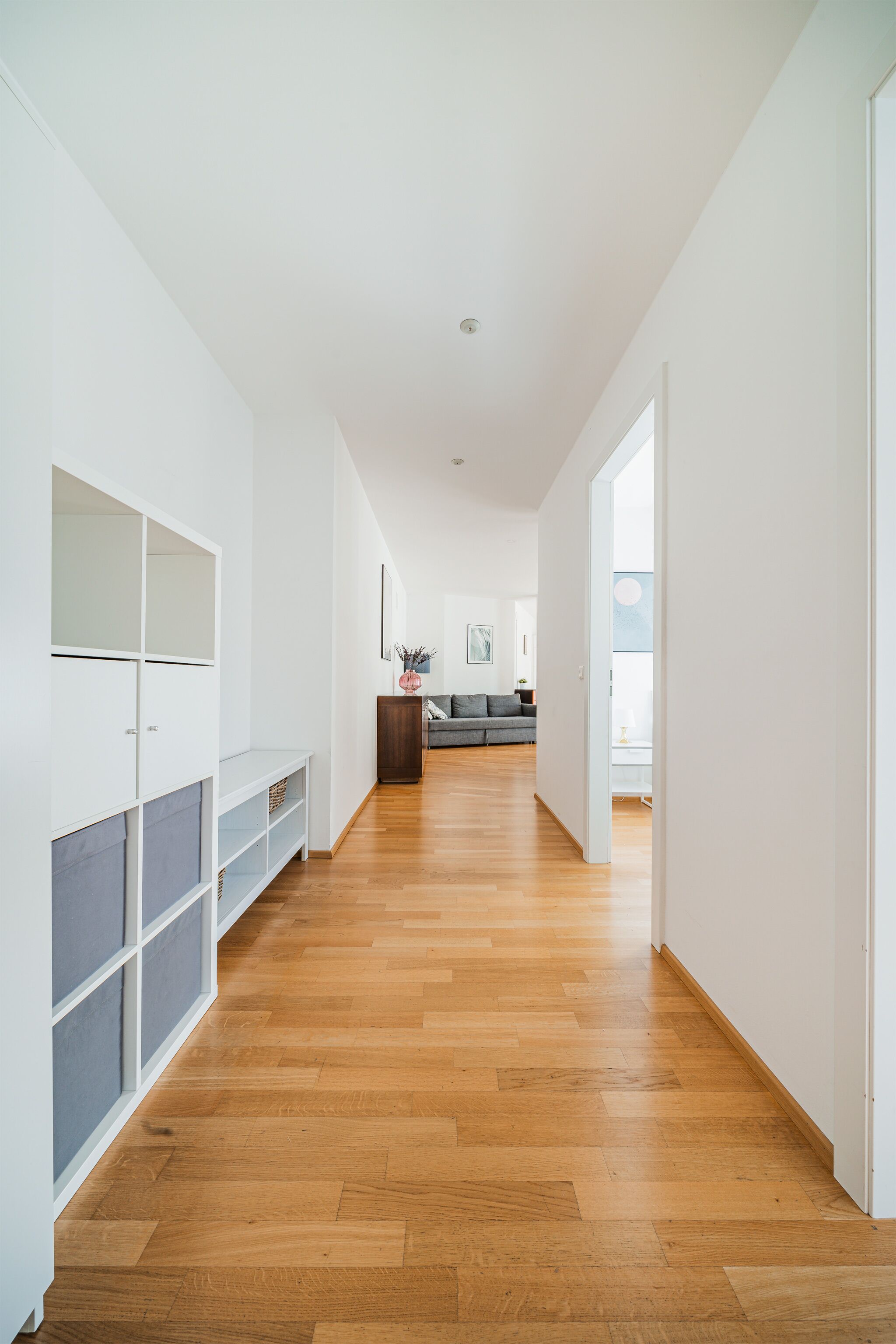 A hallway with wooden flooring, white walls, and a grey sofa in the distance.