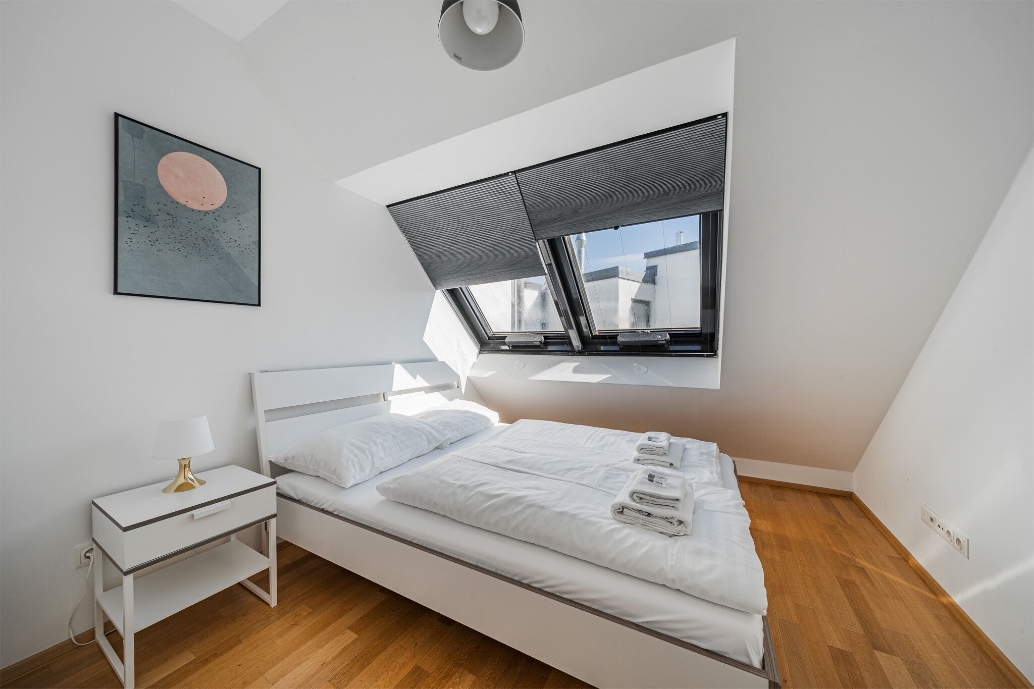 Une chambre moderne avec un puits de lumière, un lit sur lequel sont soigneusement pliées des serviettes, une table de chevet avec une lampe et une œuvre d’art accrochée au mur.