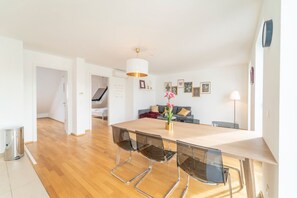 A modern dining area with a wooden table, clear glass chairs, a white pendant light, and a view of a living room with a sofa and a lamp. - easy-flat Pilgramgasse - Apartments near Metro (Vienna)
