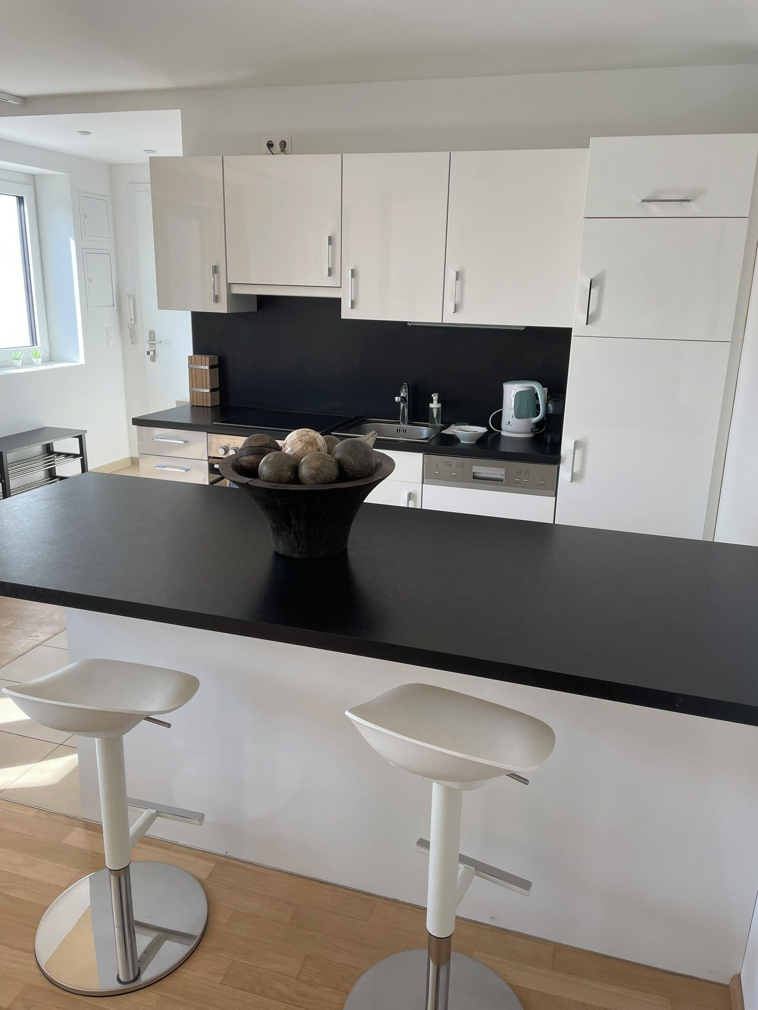 Cocina moderna con encimera negra, armarios blancos y un cuenco de fruta. Hay dos taburetes blancos en frente.
