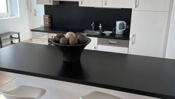 Cocina moderna con encimera negra, armarios blancos y un cuenco de fruta. Hay dos taburetes blancos en frente.