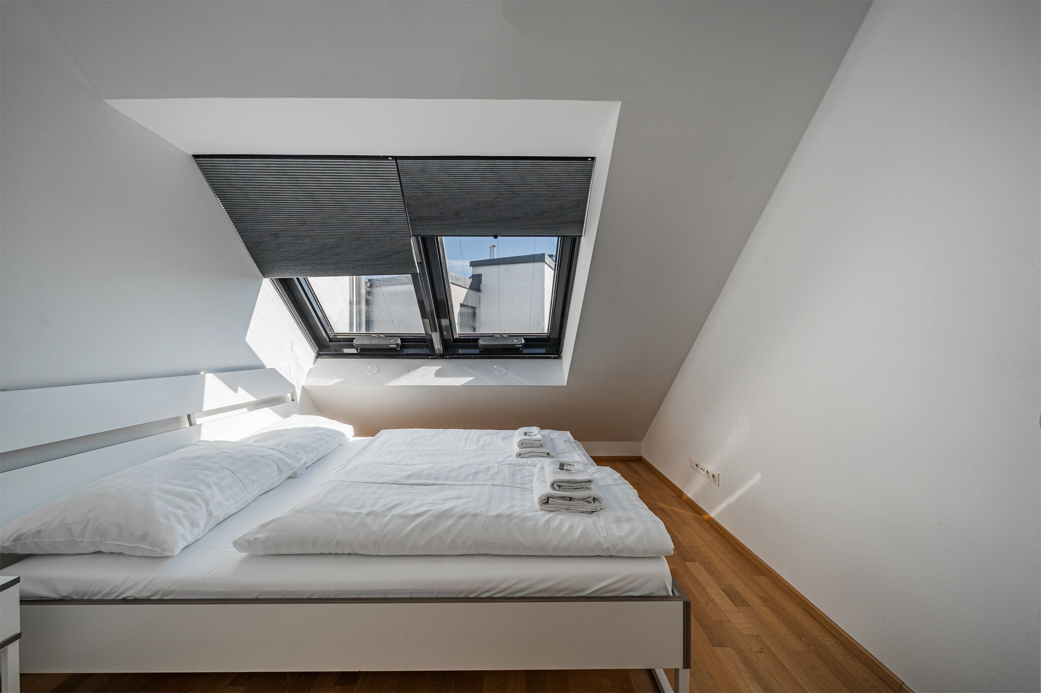A modern bedroom with a skylight, a bed with white linens, and a wooden floor.