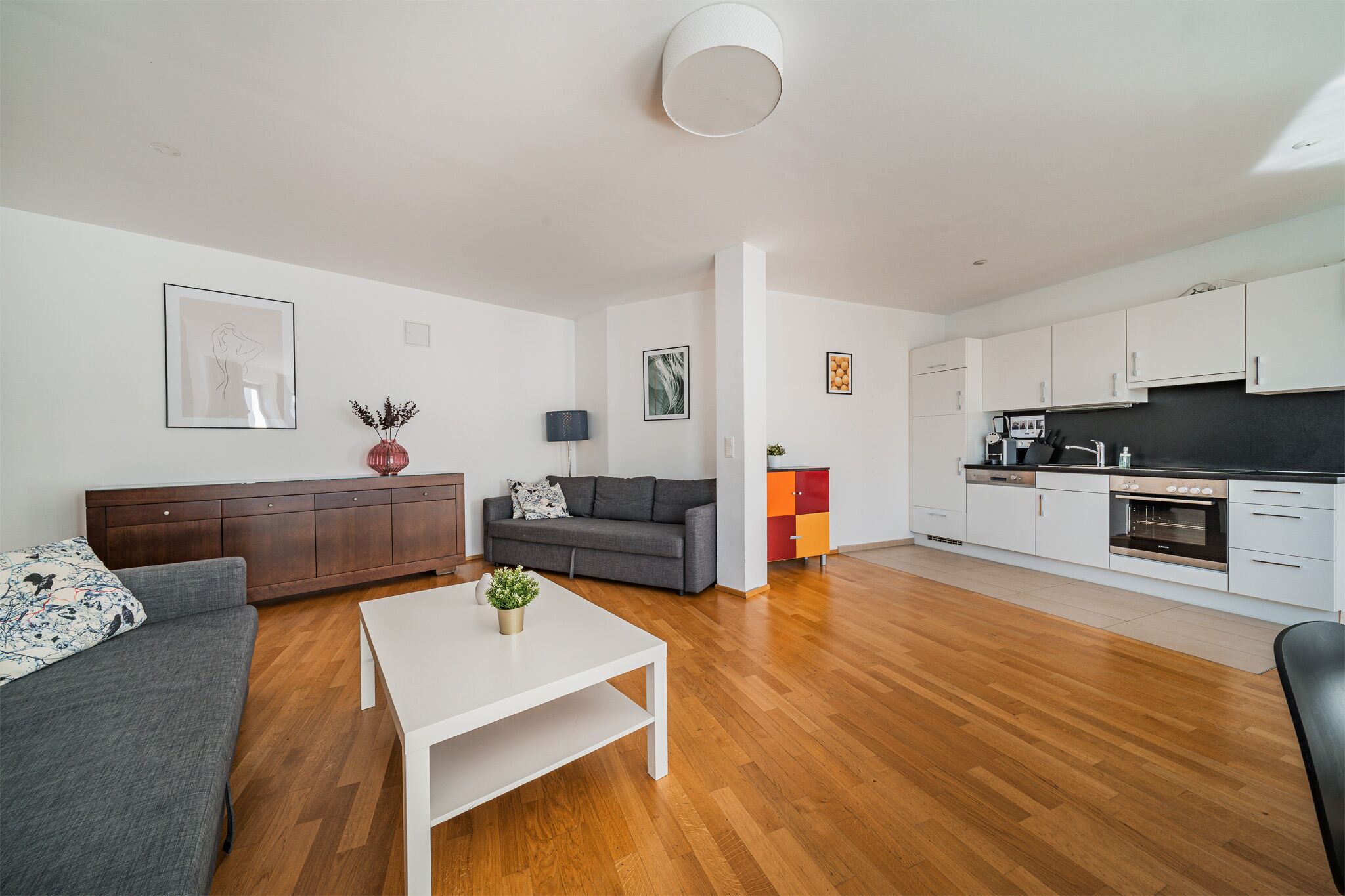 Un appartement moderne comprenant un salon avec un canapé gris, une table basse blanche et un coin cuisine doté de meubles blancs et d’un plan de travail noir.