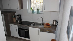 Cocina compacta con armarios blancos, un lavamanos de acero inoxidable, una cafetera y una ventana con dos macetas.
