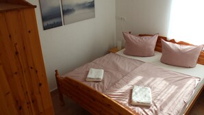 Un dormitorio con una cama de madera, dos almohadas rosas y dos toallas dobladas. Hay dos cuadros en la pared y un armario de madera.