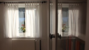 Una habitación con dos ventanas cubiertas por cortinas blancas traslúcidas, un radiador debajo de una ventana y una puerta con manija.