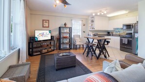 Ein Wohnzimmer mit Sofa, Hocker und einem Fernseher auf einem Schrank. Ein Essbereich mit Tisch und Stühlen ist ebenfalls vorhanden. Die Küche ist sichtbar, mit Geräten und Schränken.