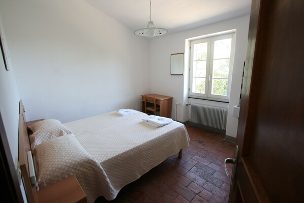 A bedroom with a bed, a wooden nightstand, a small table, a window, and a radiator. - Santuario Nostra Signora di Soviore (Monterosso al Mare)