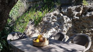 Une table surmontée d’un panier tressé contenant des fruits et une bouteille, entourée de chaises, le tout dans un décor naturel agrémenté de rochers et de végétation.