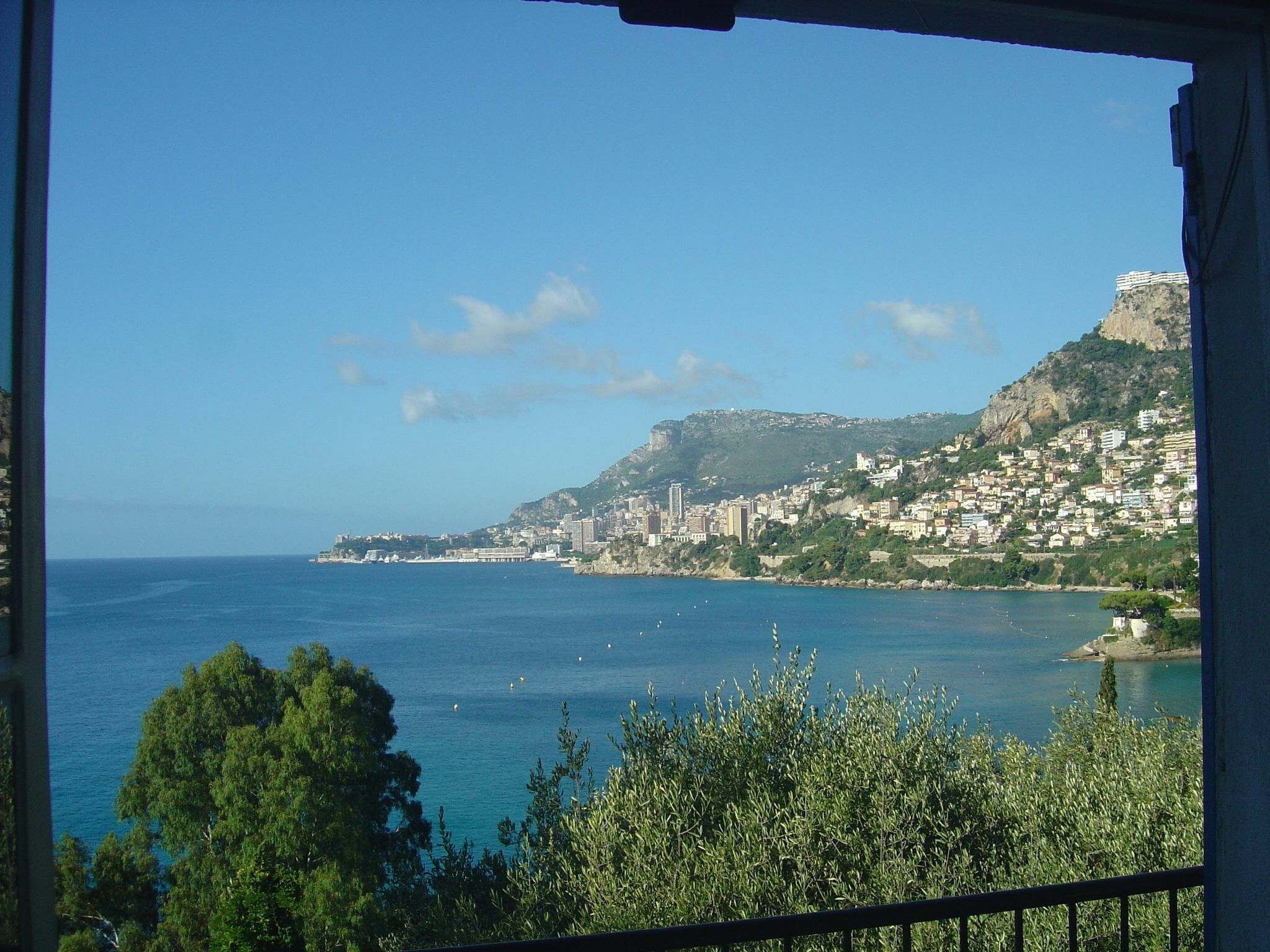 Paesaggio costiero con edifici su una collina, cielo azzurro limpido e il mare in primo piano.