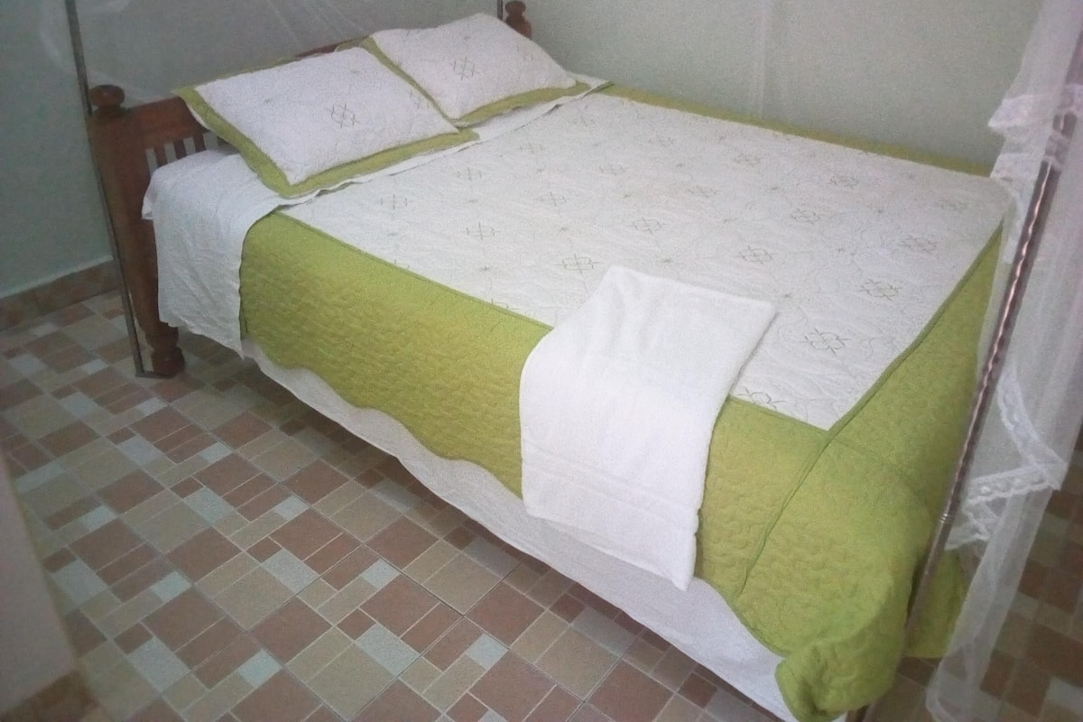 Uma cama de solteiro com edredom verde e branco, cabeceira de madeira e chão de azulejos.