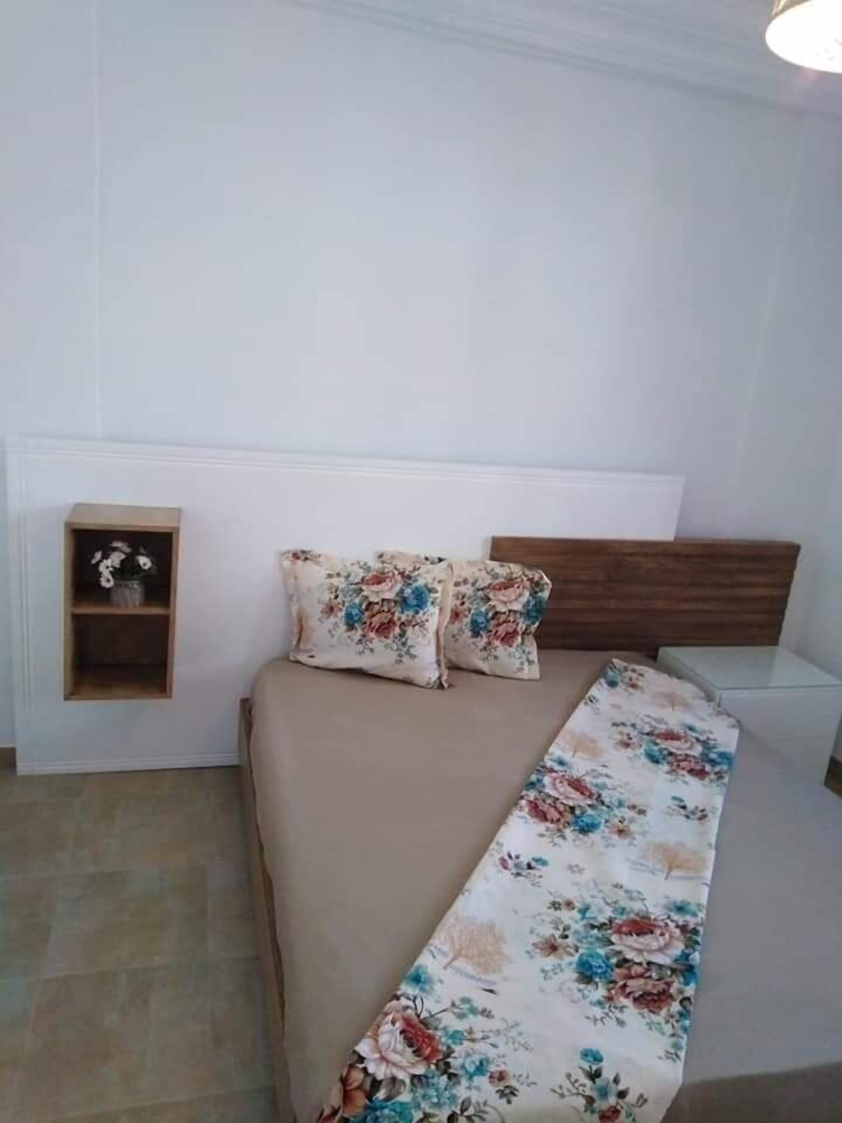 Un dormitorio con una cama, dos almohadas, una mesita de noche y un pequeño estante con un jarrón.