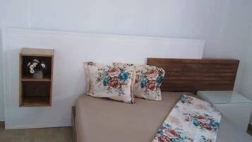 Un dormitorio con una cama, dos almohadas, una mesita de noche y un pequeño estante con un jarrón.