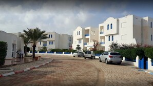 Zona residencial con edificios blancos, un camino pavimentado, coches aparcados y palmeras.