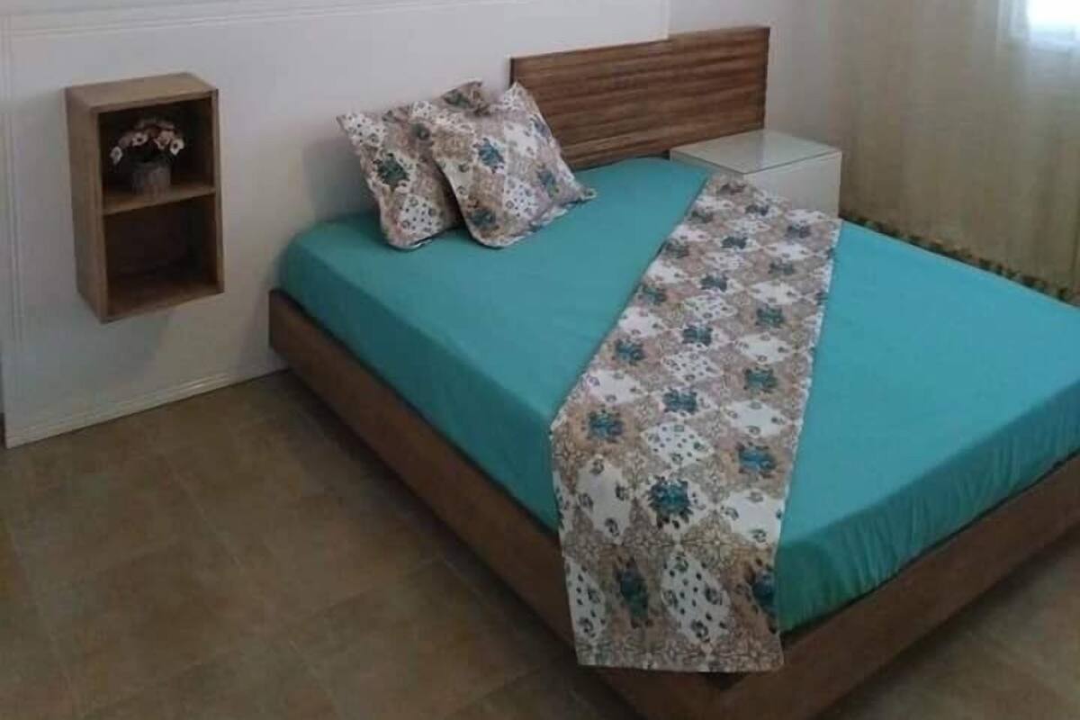 Un dormitorio con cabecera de cama de madera, ropa de cama color turquesa, un plaid floral, un pequeño estante de madera y una lámpara colgante.