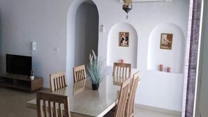 Un comedor con mesa y sillas de madera, un televisor sobre un mueble bajo y una lámpara colgante decorativa.