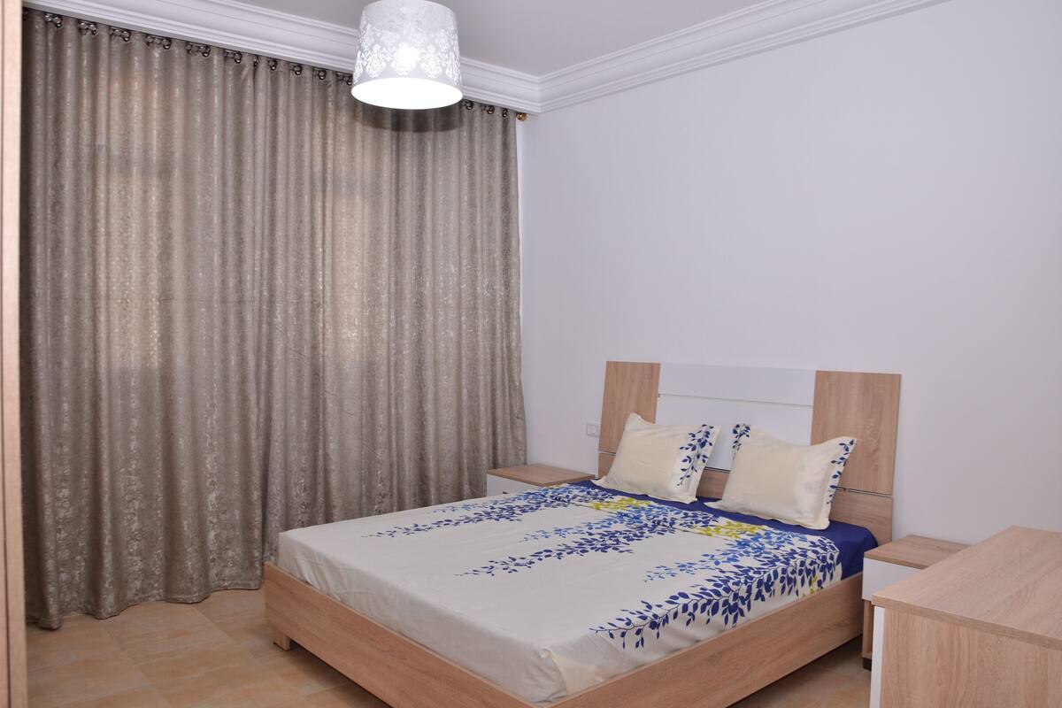 Un dormitorio con cama, cortinas, mesita de noche y mesilla.