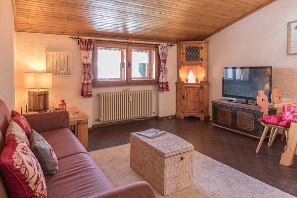 A cozy living room with a wooden ceiling, a sofa with cushions, a TV, a small table with a lamp, and a radiator.