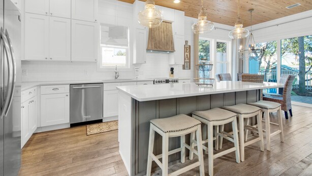 A modern kitchen with white cabinets, a stainless steel refrigerator, a built-in island with bar stools, pendant lighting, and a large window offering a view of the outdoors.