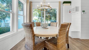A dining area with a round table, wicker chairs, a chandelier, and large windows with a view of greenery outside.