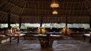 A thatched-roof lounge area with cushioned seating and tables, featuring a central wooden coffee table and hanging lamps. - Flamingo Hill Tented Camp (Nakuru)