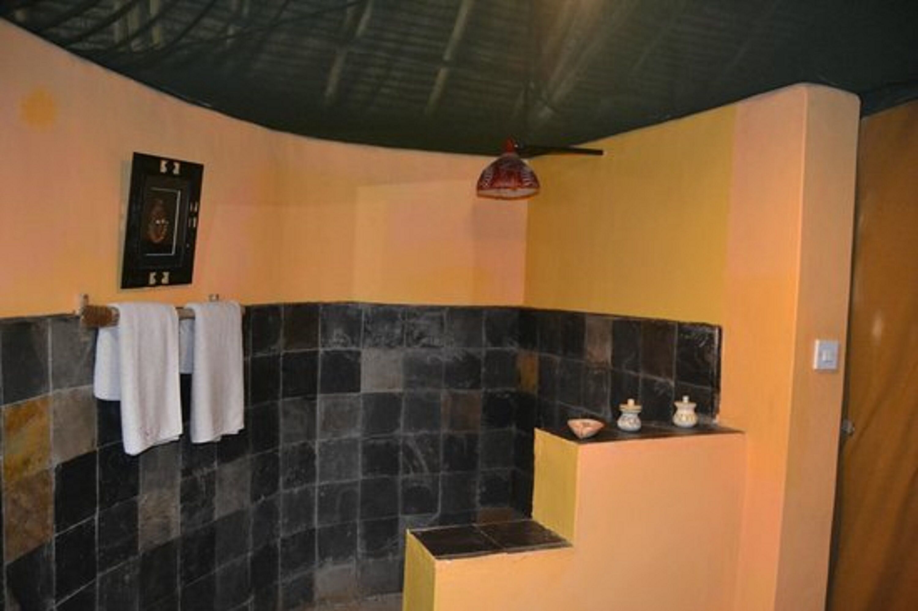 Een badkamer met een douchewand betegeld in zwart, een handdoekrek met witte handdoeken, een klein plankje met decoratieve objecten, en een plafondventilator.