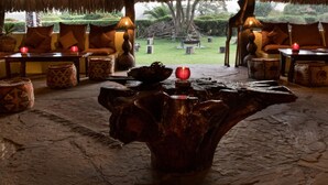A lounge area with a central wooden table, seating with cushions, and a view of a garden. - Flamingo Hill Tented Camp (Nakuru)